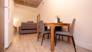 Ein moderner Essbereich mit Holztisch und vier Stühlen, einem grauen Sofa und einem Kühlschrank in einem hellen Raum mit Holzboden und einer Decke mit sichtbaren Balken.