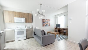 Eine moderne Küche mit Mikrowelle, Herd und Holzschränken. Im Wohnbereich befindet sich ein graues Sofa und ein kleiner Tisch. Ein farbenfrohes Gemälde hängt an der Wand.