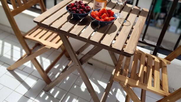A wooden table with a bottle of champagne, two bowls of fruit, and two glasses on a balcony with a railing.