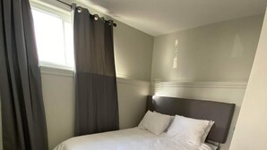 Un lit bien fait, avec une literie blanche, dans une pièce dotée d’un parquet en bois clair et d’une tête de lit sombre.