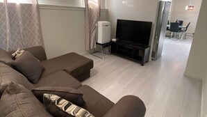 Un salon moderne avec un canapé d’angle, un téléviseur à écran plat et un coin repas à l’arrière-plan.