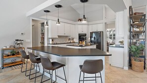 Una cocina moderna con una isla central, taburetes de bar, iluminación colgante y armarios blancos.