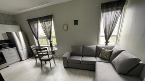 A modern living room with a grey sofa, a dining table with chairs, a kitchen area with a refrigerator and microwave, and large windows with sheer curtains. - Suite with closed bedroom (Le Convivial) (Rouyn-Noranda)