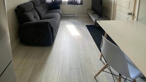 A compact living space with a sofa, a TV, a dining table, and a window with curtains. - Suite with closed bedroom (Le Convivial) (Rouyn-Noranda)