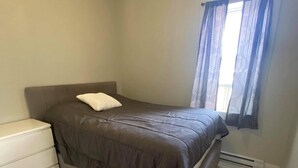 A small, neatly arranged bedroom with a bed, a bedside cabinet, and a window with curtains. - Suite with closed bedroom (Le Convivial) (Rouyn-Noranda)