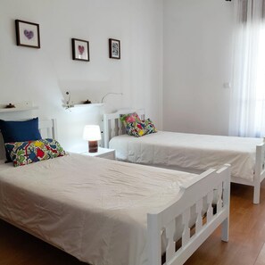 Une chambre avec deux lits blancs, une table de chevet avec une lampe et un mur orné de trois tableaux encadrés.