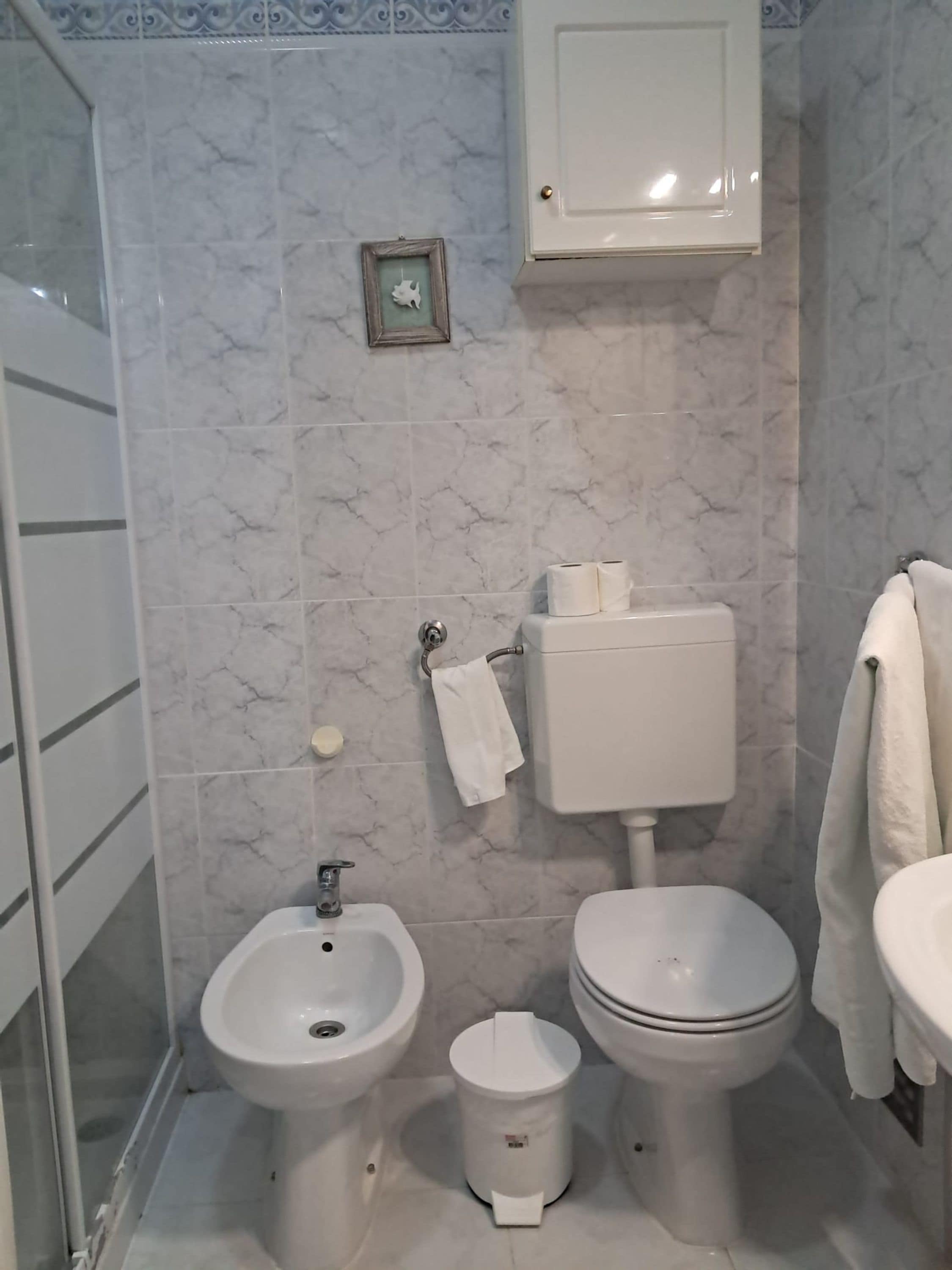 A bathroom with a bidet, toilet, and sink, all in white, with a marble-patterned wall and a small framed picture.