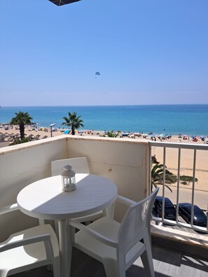 Varanda com mesa e cadeiras, com vista para uma praia com guarda-sóis e pessoas.