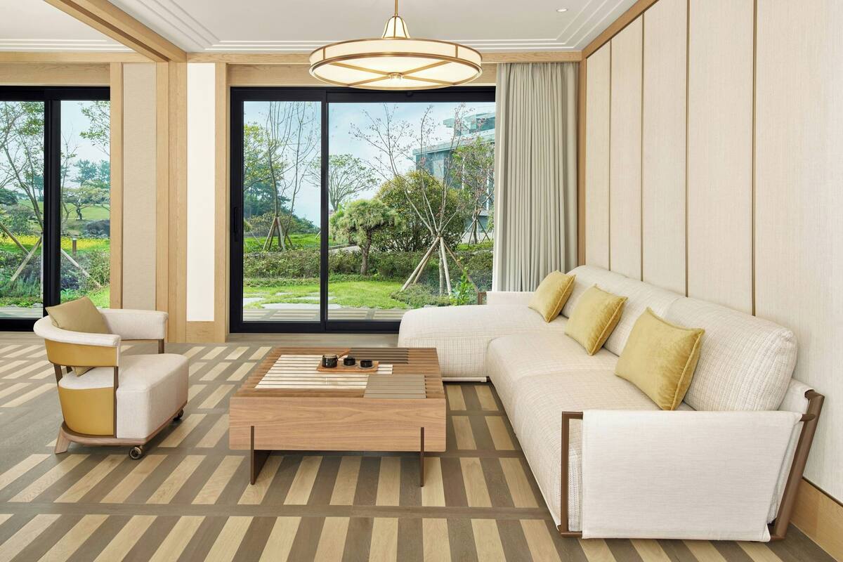 Una sala de estar moderna con sofá, sillón y mesa de centro, ventanas amplias con vistas a zonas verdes y un edificio, y un suelo estampado.