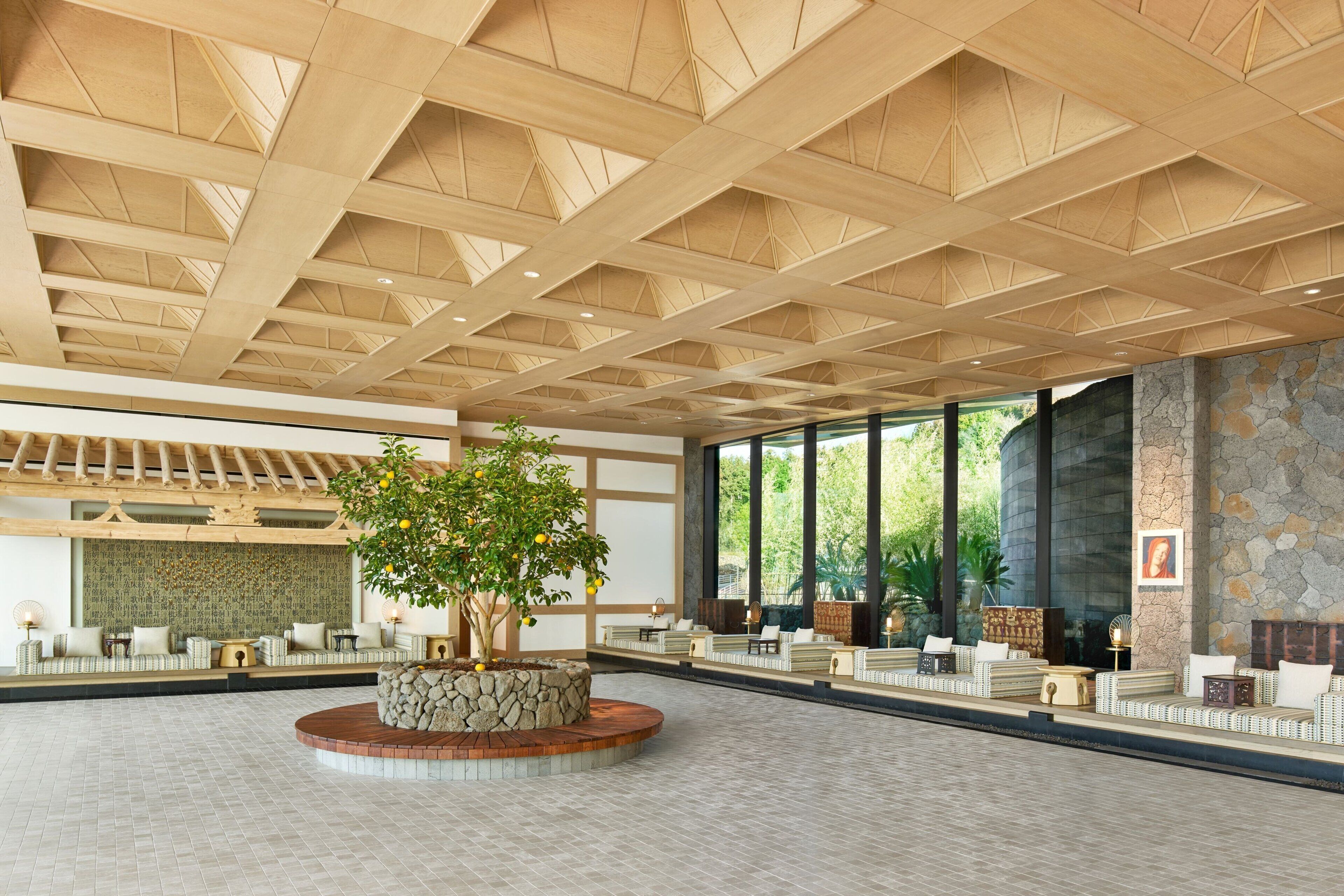 A spacious lobby with a high wooden ceiling, a central stone planter with a tree, and seating areas along the walls.