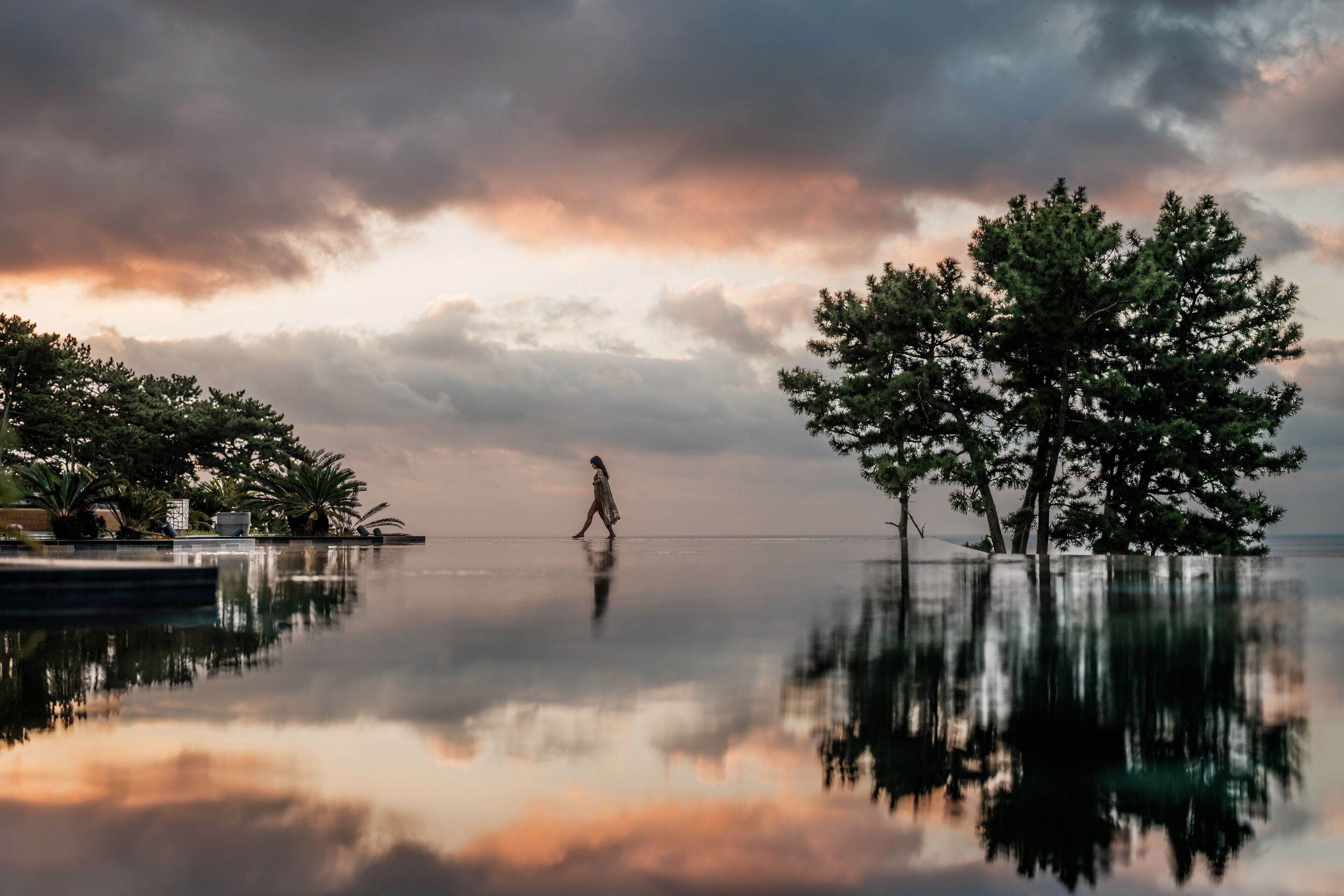 A tranquil lakeside scene with a person walking on a rope bridge, surrounded by trees and a calm body of water reflecting the sky.