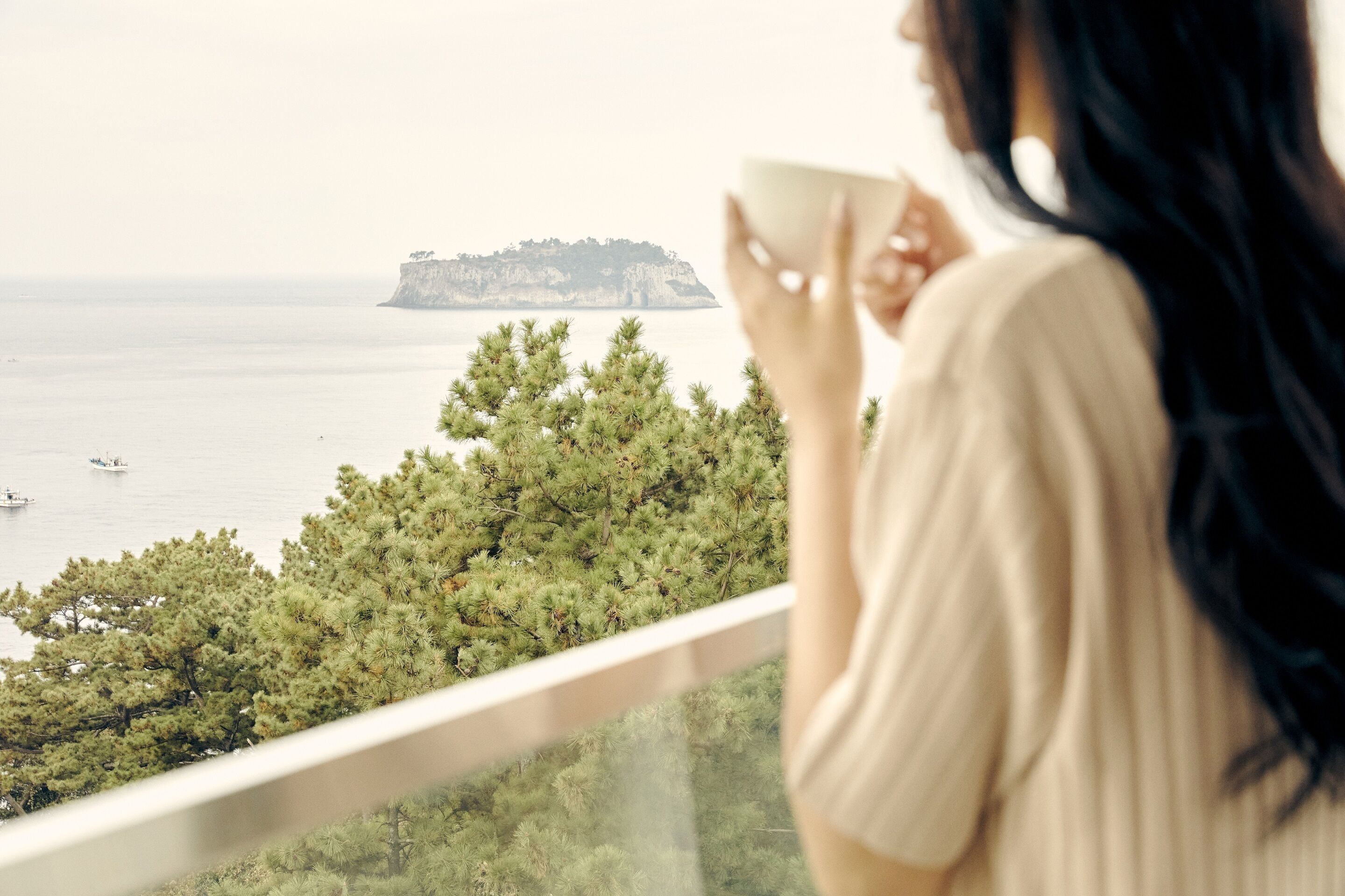 A person holding a cup, overlooking a coastal area with trees and a distant island.