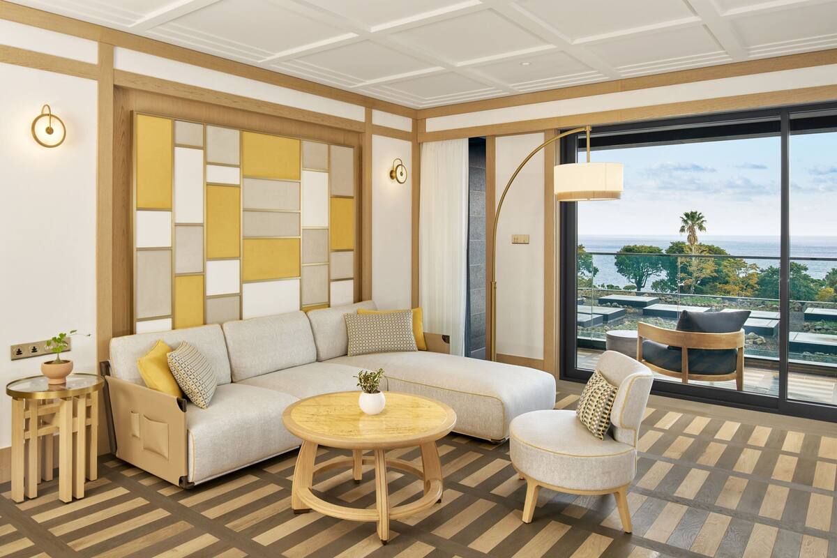 Una sala moderna con un sofá modular, una mesa de centro redonda de madera, un sillón, una mesita redonda con una planta y un ventanal con vistas al océano y palmeras.