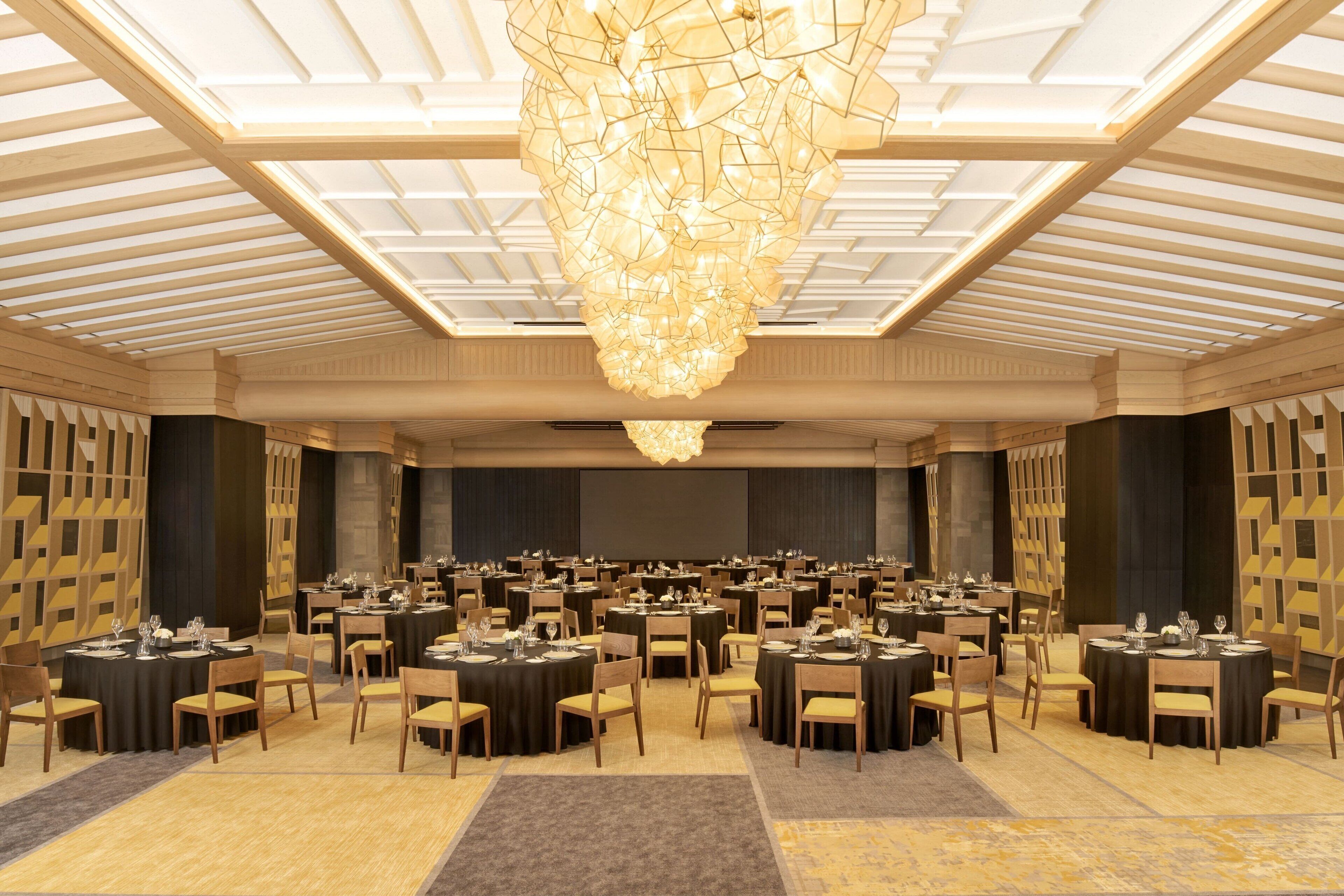A large banquet hall with round tables set for dining, a central chandelier, and a high ceiling with wooden beams.