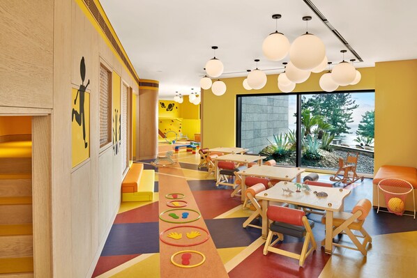 Un cuarto de juegos infantil, lleno de colores, con muebles de madera, un tapete de juegos llamativo y grandes ventanales que dan a la vista exterior.