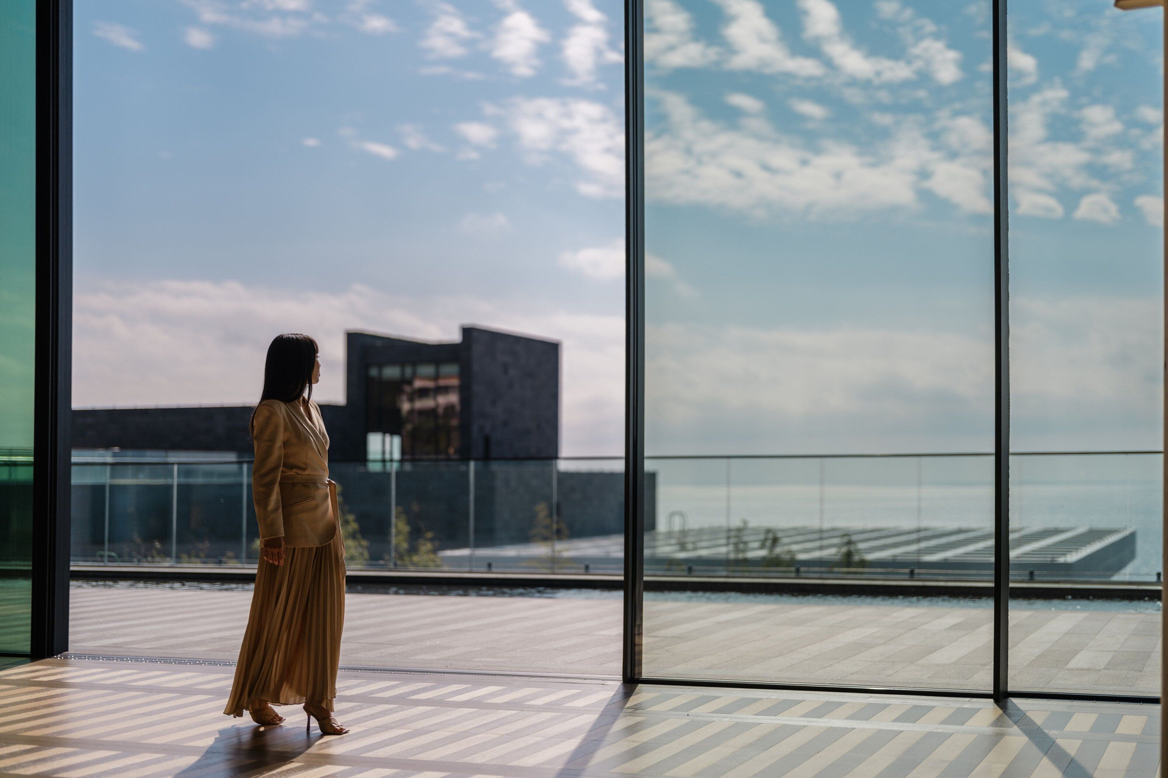 A woman in a long dress standing in a modern room with large windows overlooking a cityscape.