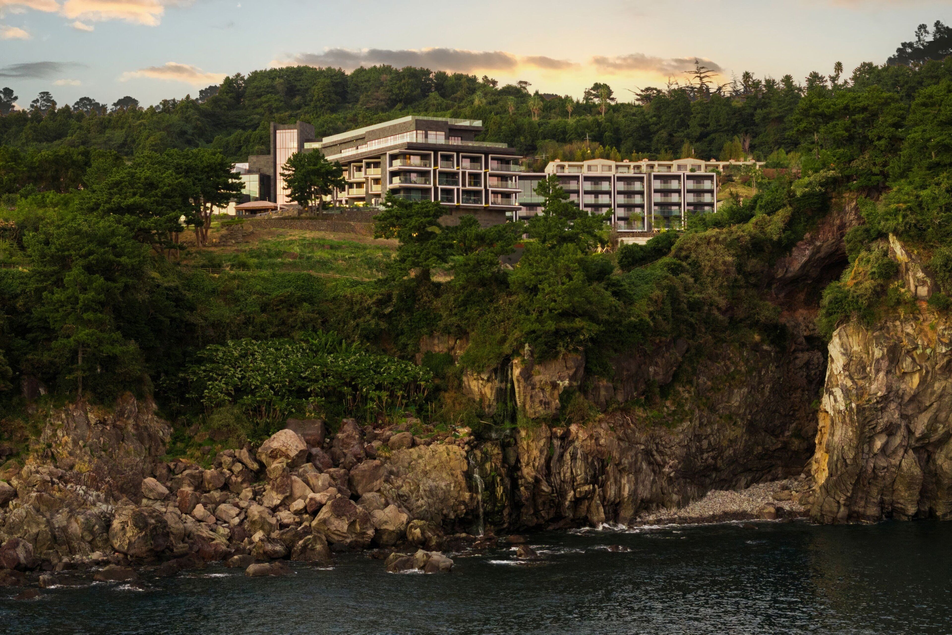 Un hotel frente al mar con varios pisos, rodeado de exuberante vegetación y acantilados rocosos.