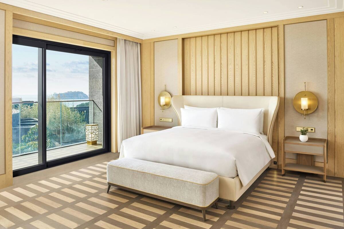Un dormitorio amplio con una cama grande, un banco y vistas a un paisaje urbano a través de un ventanal.