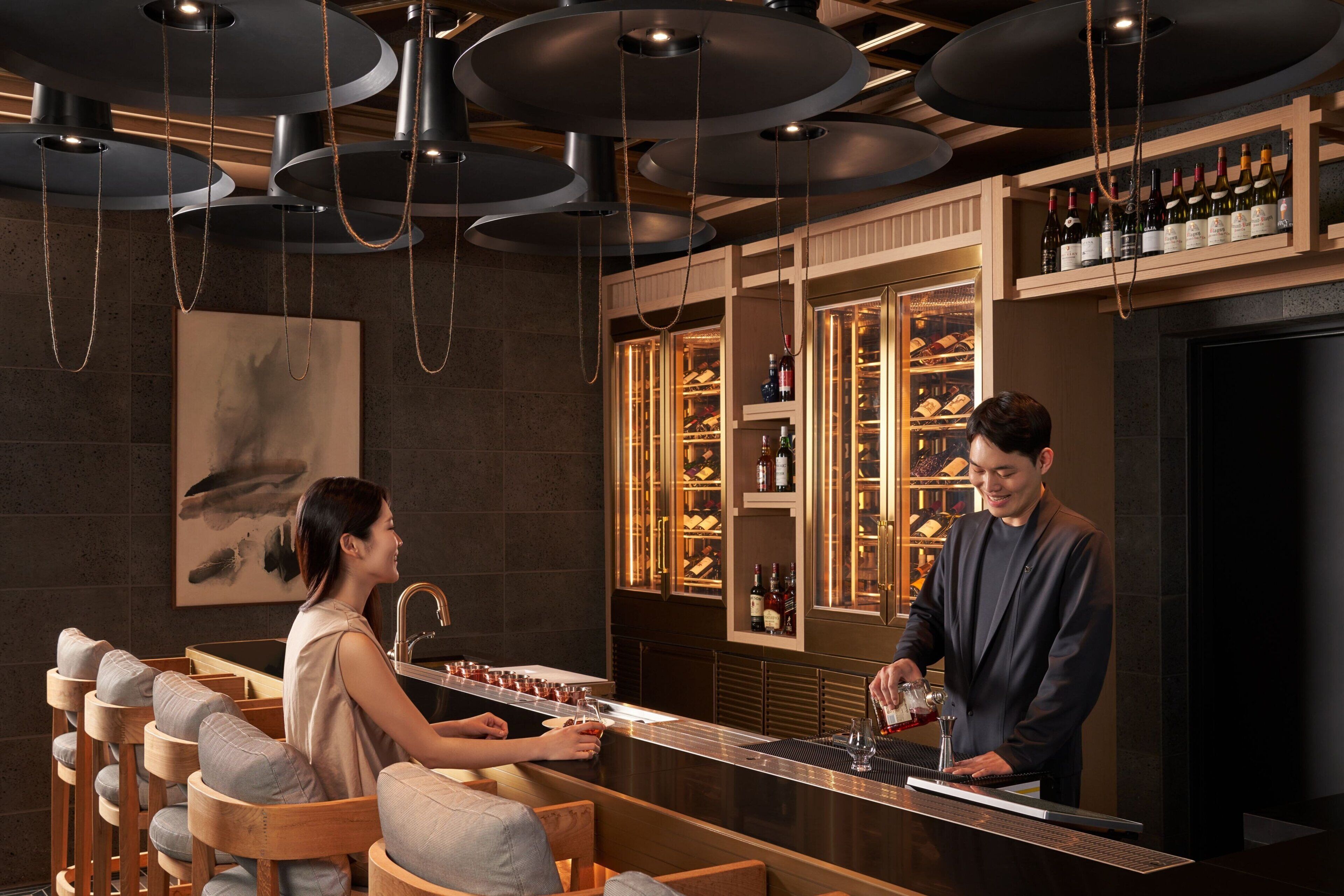 A bar area with a bartender preparing a drink, a customer at the bar, and a wine display in the background.