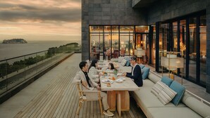 Een moderne eethoek buiten met uitzicht op zee, een houten terras en een zitgedeelte met glazen wanden.