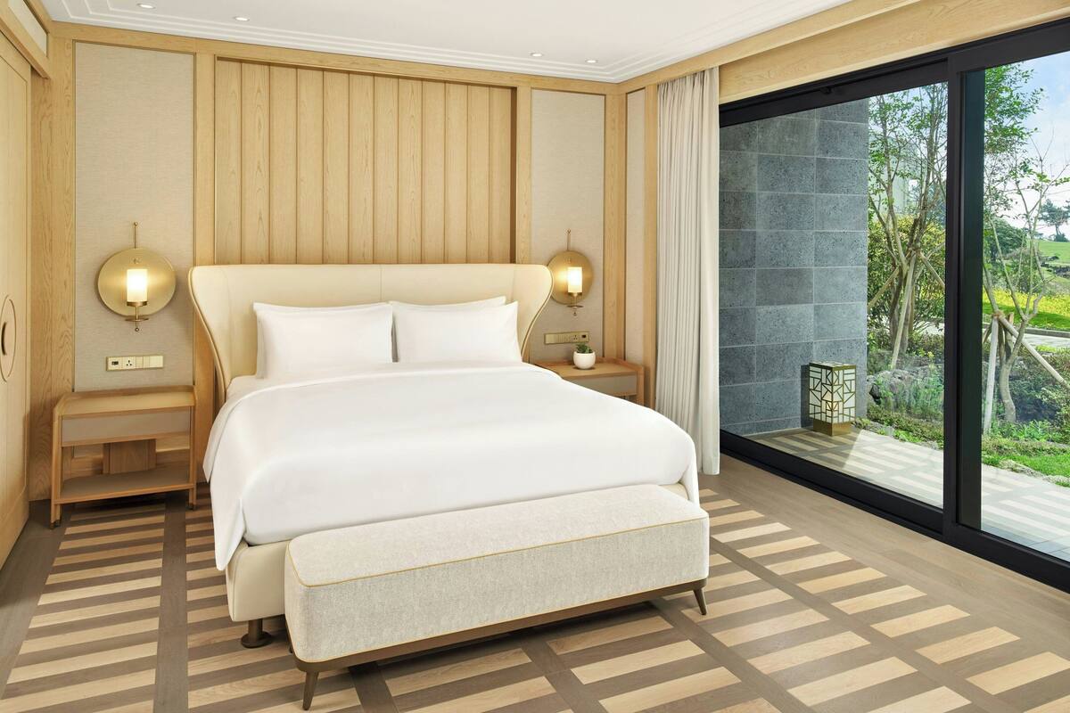 Habitación de hotel moderna con una cama grande, paneles de madera, vistas a zonas verdes y un ventanal amplio.