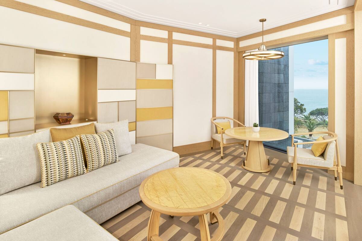 Una habitación moderna con sofá, una mesa redonda de madera y sillas, y un amplio ventanal con vista al océano.