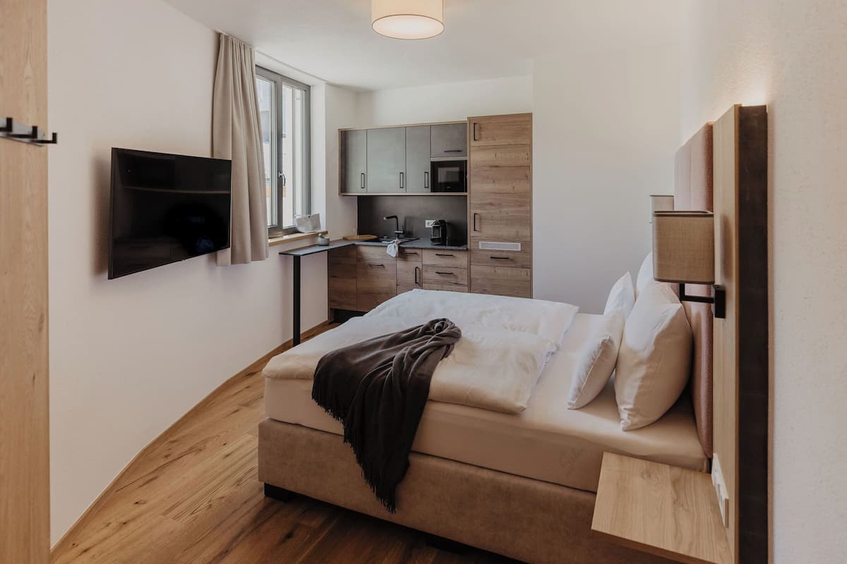 A compact room with a bed, a TV, a kitchenette, and a window with curtains.