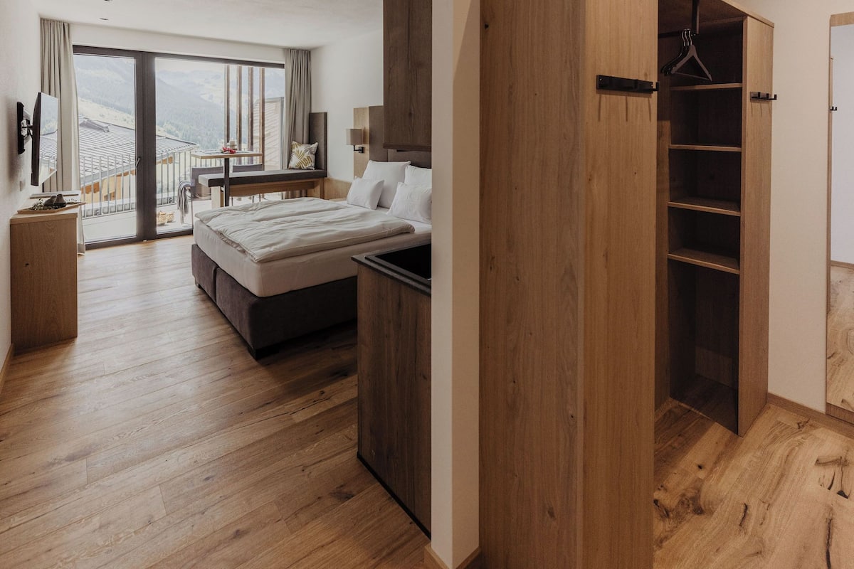 A bedroom with a bed, a wooden wardrobe, a bedside table, a desk, and a view of the mountains through a large window.