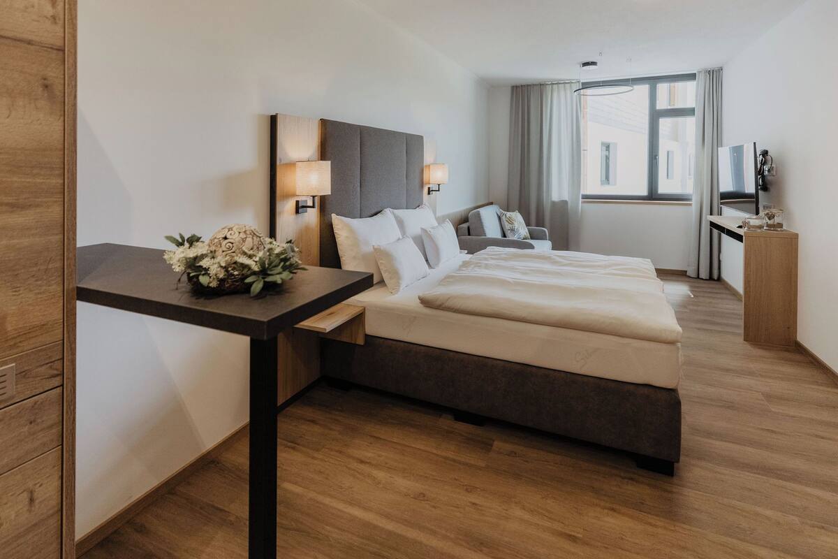 A hotel room with a bed, bedside tables, a desk, a TV, and a window with curtains.