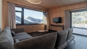Ein modernes Wohnzimmer mit einer großen Couch, einem flachbildfernseher an der Wand und Blick auf verschneite Berge durch große Fenster.