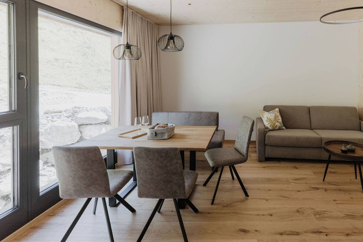A modern living room with a wooden dining table, grey upholstered chairs, a grey sofa, and a small round table with a lamp.