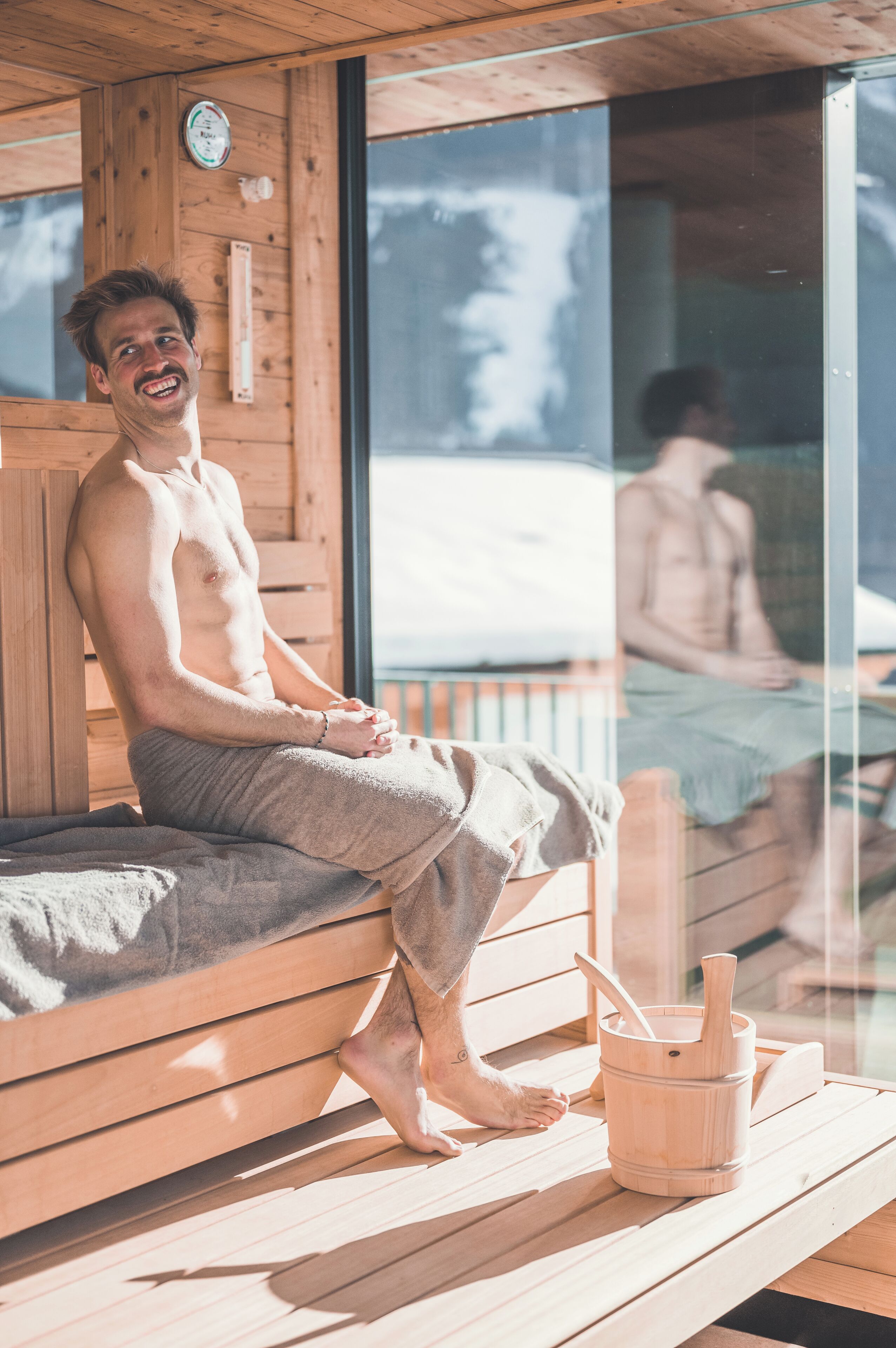 A wooden sauna with a bench, a wooden bucket, and a glass door reflecting another person.
