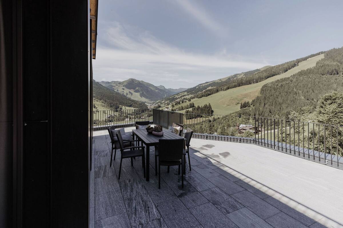 A balcony with a table and chairs overlooking a valley with mountains.