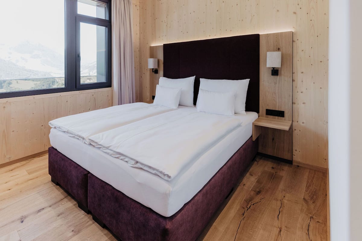 A large bed with a wooden headboard and two bedside tables, each with a lamp. The room has wooden walls and a large window with a view of mountains.