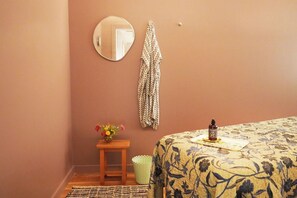 Un dormitorio con una cama, una mesita con un florero, una prenda colgada y una botella sobre la cama.