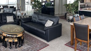 Ein geräumiges Wohnzimmer mit einer schwarzen Ledersofa, einem runden Couchtisch und einem Essbereich mit einem Marmortisch und Stühlen. Üchtis und ein großes Fenster sorgen für natürliches Licht.