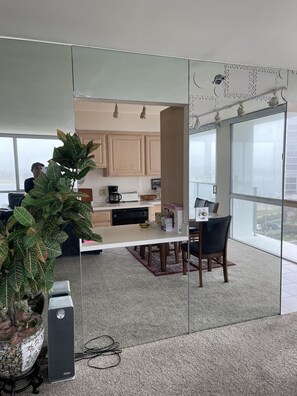 A modern office space with a glass partition, a large potted plant, a dining area with a table and chairs, and a view of the city through large windows.