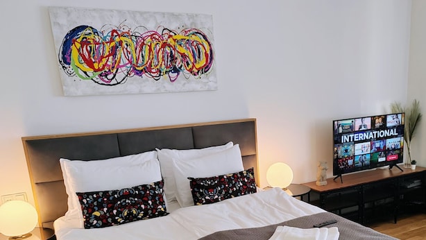 A hotel room with a large bed, two bedside tables with lamps, a TV displaying 'INTERNATIONAL', and a colorful abstract painting on the wall. - S15 Studio Apartment Zagreb Croatia-free Parking (Zagreb)
