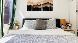 A bed with patterned bedding, a wall-mounted abstract artwork, and a window with green curtains.