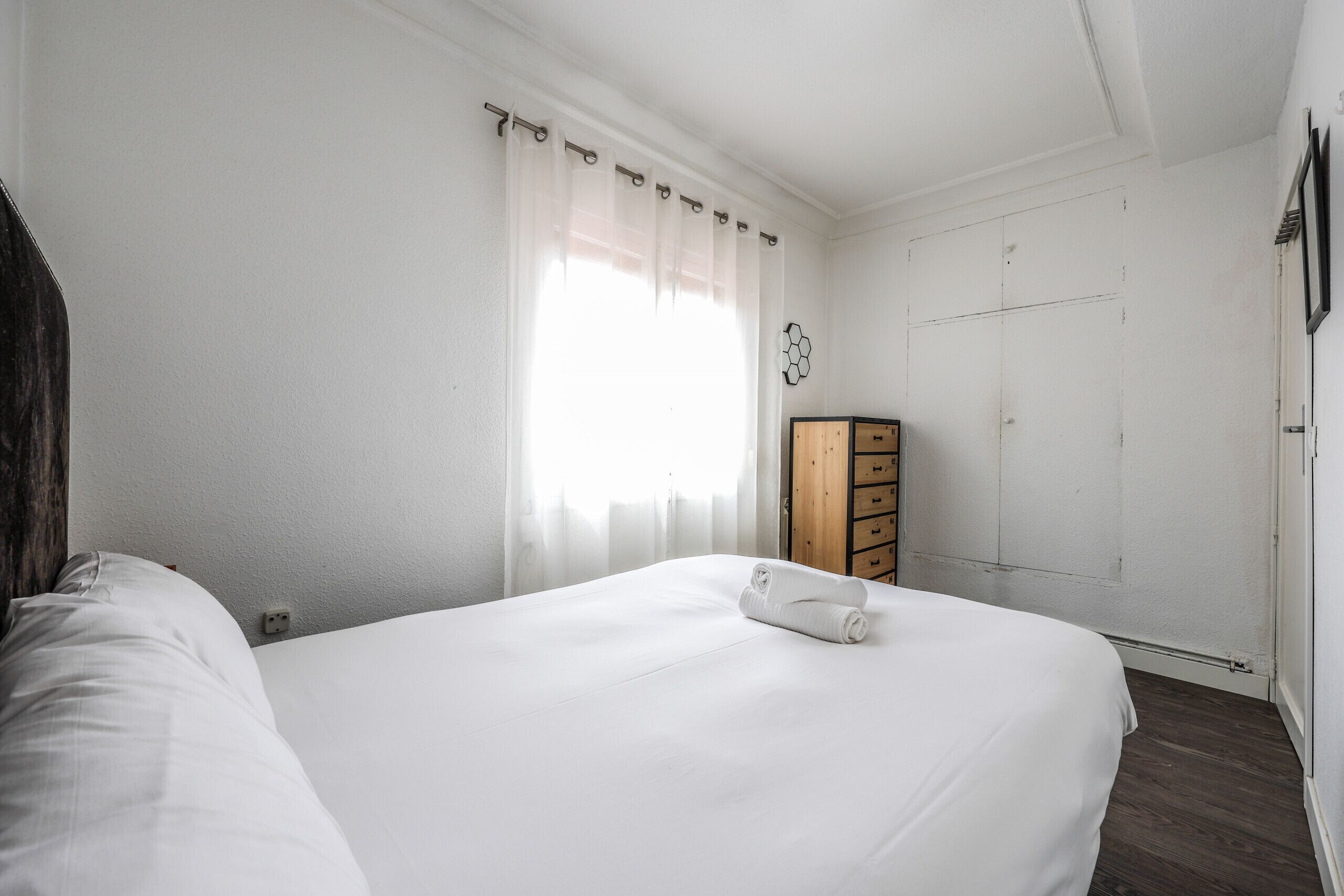Een net opgemaakt bed met witte lakens en een opgerolde handdoek, een houten garderobe en een witte deur met een honingraatdecoratie.