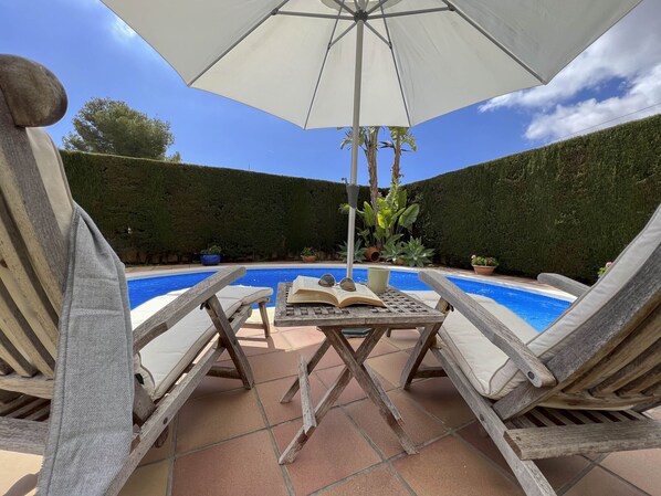 Un área junto a la piscina con sillones de descanso, una mesa con un libro abierto y un paraguas grande.
