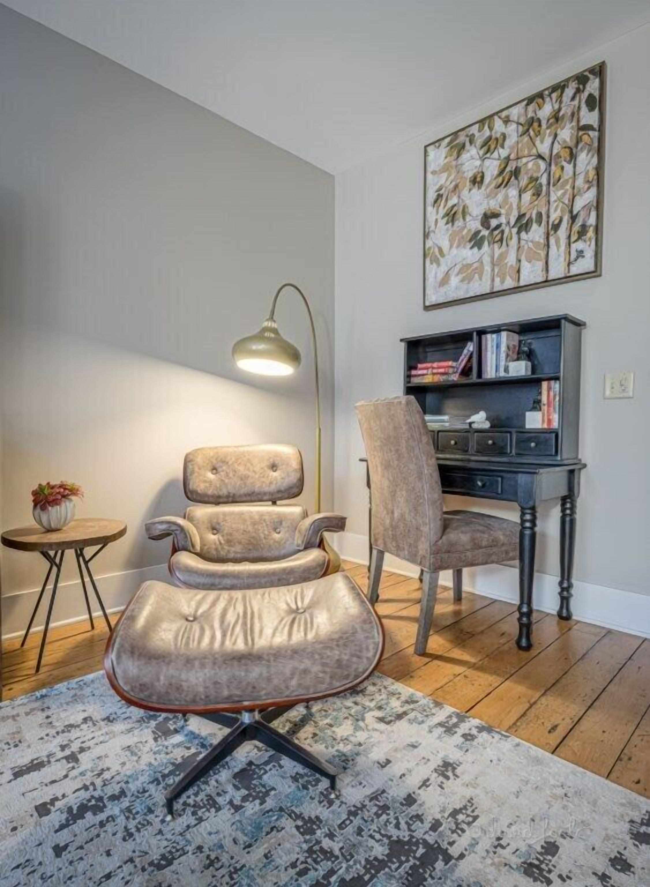 Ein moderner Bürostuhl mit hoher Rückenlehne und Armlehnen, ein Schreibtisch mit dunkler Oberfläche, eine Stehlampe, ein kleiner Beistelltisch mit einer Pflanze und ein Bücherregal an der Wand.