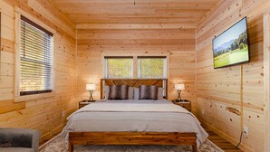 Quarto de cabana de madeira com cama, duas janelas, televisão e mesa de cabeceira com abajures.