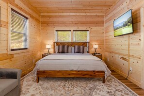 Une chambre à coucher située dans une cabane en bois, comprenant un lit, deux fenêtres, un téléviseur et une table de chevet avec des lampes.