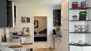 Una cocina moderna con armarios blancos, una zona de comedor con una silla blanca y una pequeña mesa. Hay una estantería con objetos decorativos y un cuadro en la pared.