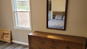 Une chambre avec une commode en bois, un grand miroir, un lit recouvert d’une couverture marron et une fenêtre donnant sur des arbres et une clôture.