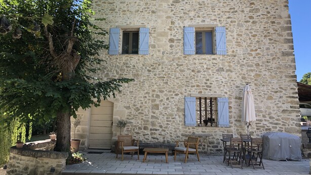 A stone building with multiple windows and shutters, outdoor seating area with tables and chairs, and a tree in the foreground. - New! Renovated cottage in old mill with swimming pool (Forcalquier)