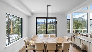 Un comedor moderno con una mesa redonda grande, seis sillas y ventanas amplias con vistas a árboles y montañas.