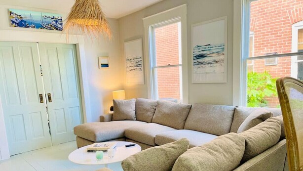 A living room with a beige sectional sofa, a round coffee table, and a lamp. There are two windows, a door, and artwork on the walls. - Salt Box II, One Block to the Beach (Cape Charles)