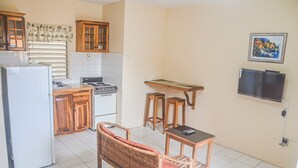 Cocina compacta con refrigerador, estufa y alacenas de madera. Hay un comedor con una silla de mimbre y una pequeña mesa. Un televisor está montado en la pared.